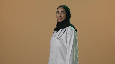 مفهوم الطب والرعاية الصحية، بورتريه لطبيبة عربية خليجية سعودية محجبة ترتدي العباءة مع المعطف الابيض تقف بأيدي متقاطعة بايماءات الثقة بالنفس، مهنة ووظيفة نسائية، تقديم الخدمات الصحية للمرضى، العمل في القطاع الصحي في المملكة، خلفيه بنية