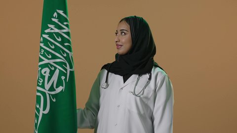 مفهوم حب الوطن و الانتماء الية، ذكرى توحيد المملكة العربية السعودية، الاحتفال باليوم الوطني السعودي بفخر وعزة ، العلم السعودي راية التوحيد والاسلام , بورتريه لطبيبة عربية خليجية سعودية محجبة ترتدي المعطف الطبي تقف باستقامة تنظر بايماءات الفخر، خلفية بنية