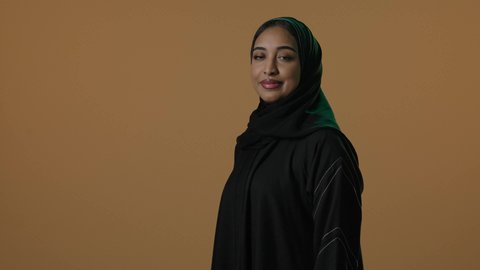 بورتريه مقرب لامرأة عربية خليجية سعودية مبتسمة ترتدي العباءة السوداء والحجاب، الوقوف بايدى متقاطعة، ايماءات الفخر والاعتزاز، النظرالى الكاميرا، خلفية بنية