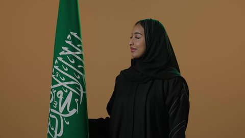 Celebrating the anniversary of the Saudi National Day, Saudi Flag Day on March 11, a portrait of a smiling Arab Gulf Saudi woman wearing a black abaya and hijab holding the Saudi flag, the National Day on the 8th of Rabi' al-Awwal, brown background.