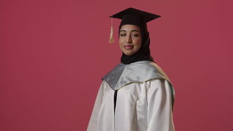 البهجة والسعادة بالوصول لحلم النجاح والتفوق، بورتريه لخريجة عربية سعودية خليجية محجبة ترتدي ثوب التخرج الابيض والقبعة تقف بأيدي متقاطعة تنظر إلى الكاميرا بتعابير النجاح، الاحتفال بإنهاء الدراسة الجامعية، حفلة التخرج من الجامعة، خلفية حمراء