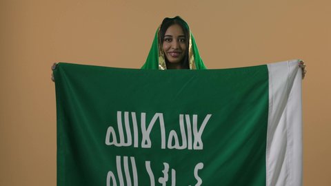 ارتداء الزي الشعبي للمنطقة الوسطى في مناسبة وطنية، بورتريه مقرب لامرأة عربية خليجية سعودية ترتدي الشيلة والبشت الاخضر تحمل راية وشعار يوم التأسيس بتعابير الفخروالاعتزاز، ذكرى تأسيس الدولة السعودية الأولى 22 فبراير، خلفية بنية