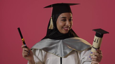 حفل التخرج الجامعي، النظر الى الكاميرا بايماءات البهجة والسعادة بالوصول لحلم النجاح، بورتريه مقرب لخريجة عربية خليجية سعودية محجبة مبتسمة ترتدي العباءة وقبعة التخرج تمسك شهادة التفوق وترفع قبضة اليد بتعابير الفوز وتحقيق الاهداف، خلفية حمراء