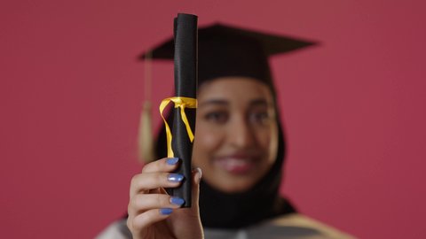 بورتريه مقرب ليد خريجة عربية خليجية سعودية محجبة ترتدي زي التخرج والقبعة تحمل بيدها الشهادة، خلفية حمراء، الاحتفال بإنهاء الدراسة الجامعية، النجاح والتفوق والتخرج من الجامعة، إيماءات السعادة بالوصول الى الحلم، النظر الى الكاميرا بايماءات الفخر