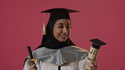 الاحتفال بإنهاء الدراسة الجامعية، النجاح والتفوق والتخرج من الجامعة، إيماءات السعادة بالوصول الى الحلم، النظر الى الكاميرا بايماءات الفخر، بورتريه لخريجة عربية خليجية سعودية محجبة ترتدي زي التخرج والقبعة تحمل بيدها الشهادة، خلفية حمراء