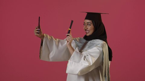 A side portrait of a Saudi Arabian Gulf female graduate wearing a graduation gown and cap, holding a diploma while making a video call on her mobile phone, using modern technology, sharing the joy of achieving the dream of success with family and friends, with a red background.