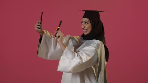 مشاركة الأهل والأصدقاء فرحة الوصول لحلم النجاح، بورتريه من الجانب لخريجة عربية خليجية سعودية محجبة ترتدي زي التخرج والقبعة تمسك الشهاده تقوم بإجراء مكالمة فيديو عبر الهاتف المحمول،استخدام جهاز تقني حديث، خلفية حمراء