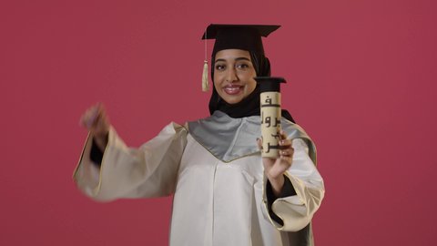 النجاح والتفوق والتخرج من الجامعة، إيماءات السعادة بالوصول الى الحلم، الاحتفال بإنهاء الدراسة الجامعية، النظر الى الكاميرا بايماءات الفخر، بورتريه لخريجة عربية خليجية سعودية محجبة ترتدي زي التخرج والقبعة تحمل بيدها الشهادة، خلفية حمراء
