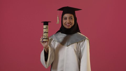 إيماءات السعادة بالوصول الى الحلم، النجاح والتفوق والتخرج من الجامعة، الاحتفال بإنهاء الدراسة الجامعية، النظر الى الكاميرا بايماءات الفخر، بورتريه لخريجة عربية خليجية سعودية محجبة ترتدي زي التخرج والقبعة تحمل بيدها الشهادة، خلفية حمراء