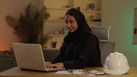 مواكبة التطورات والتكنولوجيا الحديثة في المملكة العربية السعودية، مفهوم ادارة المشاريع وتدقيقها، مهندسة عربية خليجية سعودية محجبة ترتدي العباءة السوداء بجانبها خوذة تقوم بالعمل على اللابتوب، متابعة المشاريع الإنشائية