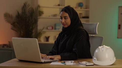 The use of laptops in the engineering sector, the concept of project management and auditing, a veiled Arab Gulf Saudi engineer wearing a black abaya next to a helmet working on a laptop, monitoring construction projects.
