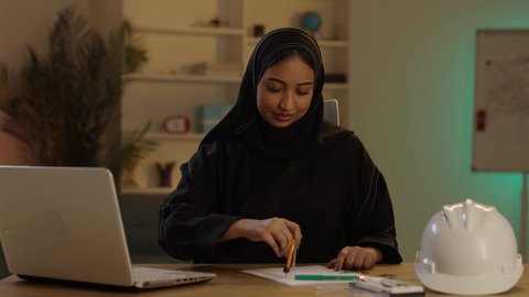 Using engineering tools and equipment on an engineering design plan, a Saudi Gulf Arab engineer wearing a black abaya and a hijab is drawing next to a computer, monitoring construction projects.