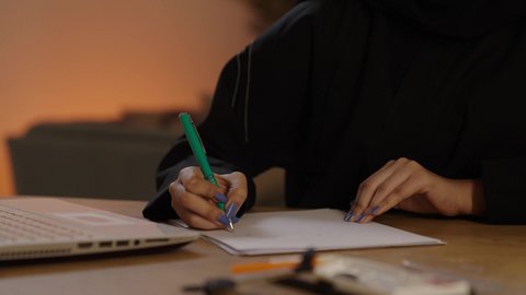 اعداد تقارير ومهام العمل و دراسة جدوى المشاريع والأعمال, التخطيط للعمل ومراجعة نتائج الاجتماع، شابة سعودية تقوم بانجاز المهام، تصوير مقرب ليد امرأة عربية خليجية سعودية ترتدي العباءة السوداء تجلس على المكتب تمسك بيدها القلم