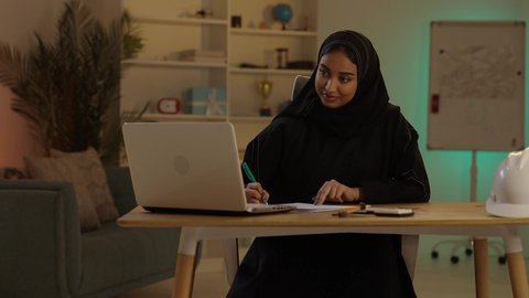متابعة استراتيجات المشروع، مهندسة عربية خليجية سعودية محجبة ترتدي العباءة السوداء تقوم بالعمل على التابلت و تدوين الملاحظات بإيماءات الاندماج والتركيز، إنجازالمهام اليومية في المكتب بجانب خوذتها, مهن ووظائف نسائية سعودية
