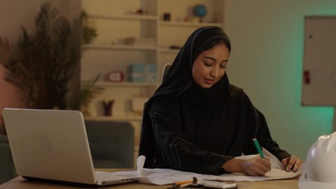 الاهتمام بالتطور العمراني، تدوين الملاحظات على مخطط تصميم هندسي، مهندسة عربية خليجية سعودية محجبة ترتدي العباءة السوداء تقوم بالعمل على التابلت بإيماءات الاندماج و التركيز، إنجاز المهام اليومية في المكتب بجانب خوذتها