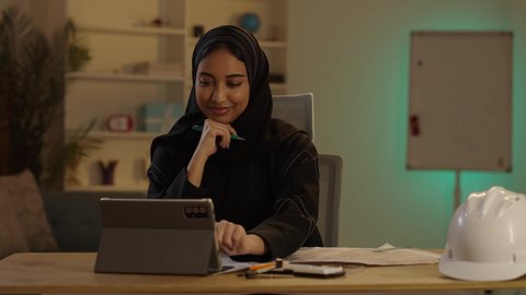 تدوين الملاحظات على مخطط تصميم هندسي، مهندسة عربية خليجية سعودية محجبة ترتدي العباءة السوداء تقوم بالعمل على التابلت بإيماءات الاندماج والتركيز، استخدام الوسائل التقنية الحديثة، إنجازالمهام اليومية في المكتب بجانب خوذتها