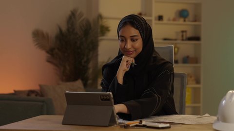 مهندسة عربية سعودية خليجية محجبة ترتدي العباءة السوداء تقوم بتدوين الملاحظات على مخطط تصميم هندسي، استخدام الوسائل التقنية الحديثة، العمل على جهازالتابلت، إنجازالمهام اليومية في المكتب بجانب خوذتها, مهن ووظائف نسائية سعودية