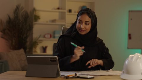 استخدام الوسائل التقنية الحديثة، مهندسة عربية سعودية خليجية محجبة ترتدي العباءة السوداء تقوم بتدوين الملاحظات على مخطط تصميم هندسي، العمل على جهاز التابلت، إنجاز المهام اليومية في المكتب بجانب خوذتها, مهن ووظائف نسائية سعودية