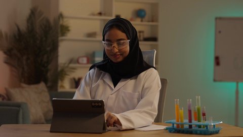 دمج التقنية الحديثة والمتطورة بالمجال الطبي, إجراء التجارب والبحوث العلمية, مختبر التحاليل والفحوصات الطبية، طبيبة صيدلانية عربية خليجية سعودية محجبة ترتدي المعطف الطبي الأبيض و نظارات الحماية تقوم بالنقرعلى جهازالتابلت، مهن ووظائف نسائية