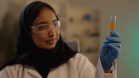 مختبر التحاليل والفحوصات الطبية، إجراء التجارب الكيميائية باستخدام أنابيب الزجاجية المعقمة, طبيبة صيدلانية عربية خليجية سعودية محجبة ترتدي المعطف الطبي الأبيض ونظارات الحماية والقفازات الطبية تمسك أنبوب الاختبار، مهن ووظائف نسائية