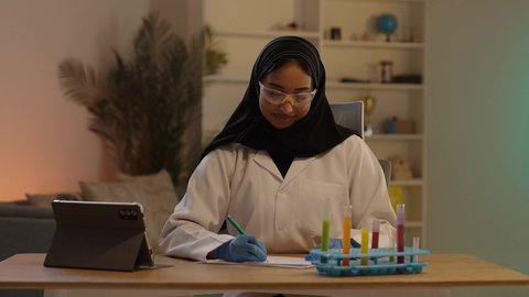 مفهوم الصيدلة والرعاية الصحية, الادوية والمستلزمات الطبية، طبيبة صيدلانية عربية خليجية سعودية محجبة ترتدي المعطف الطبي الأبيض و نظارات الحماية و القفازات الطبية تقوم بكتابة الوصفات العلاجية و بجانبها جهازا لتابلت، مهن ووظائف نسائية