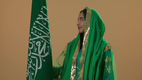 السعادة بالانتماء للوطن، اليوم الوطني السعودي 23 سبتمبر، يوم العلم 11 مارس، ارتداء الزي الشعبي للمنطقة الوسطى في مناسبة وطنية, يوم بدينا 22 فبراير, بورتريه مقرب لامرأة عربية خليجية سعودية ترتدي الشيلة والبشت الاخضر تحمل علم المملكة، خلفية بنية