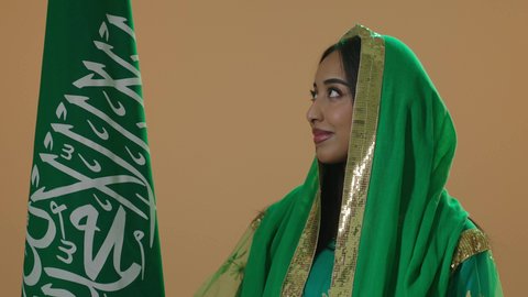 راية التوحيد وسيف القوة، اليوم الوطني السعودي 23 سبتمبر، يوم العلم 11 مارس، ارتداء الزي الشعبي للمنطقة الوسطى في مناسبة وطنية, يوم بدينا 22 فبراير, بورتريه مقرب لامرأة عربية خليجية سعودية ترتدي الشيلة و البشت الاخضرتحمل علم المملكة، خلفية بنية