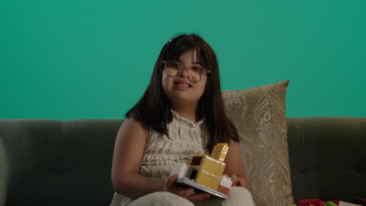Looking at the camera with expressions of happiness and joy, a portrait of a Saudi girl with Down syndrome sitting on the couch assembling blocks, caring for and developing children's skills, a recreational activity for people with special needs, having a good time at home, green background.