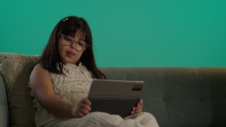 دمج ذوي الاحتياجات الخاصه في الحياه اليومية، بورتريه لطفلة سعودية مصابة بمتلازمه داون ترتدى النظارة تجلس على الاريكة تمسك التابلت، استخدام التكنولوجيا والأجهزة التقنية الحديثة، خلفية حضراء