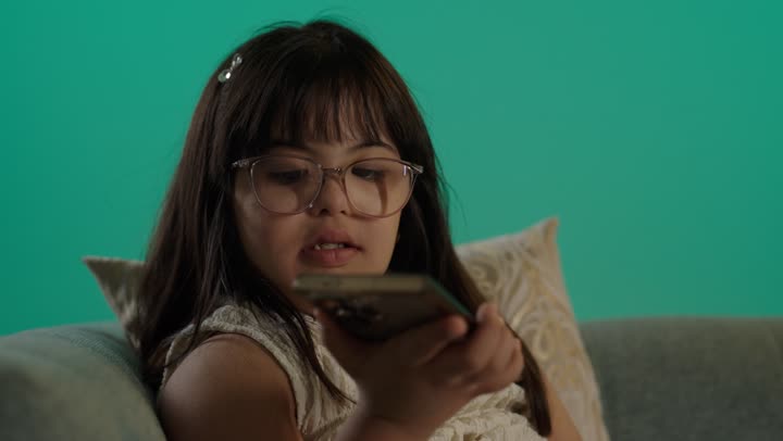 تعابير وايماءات الاندماج والسرور، بورتريه لطفلة سعودية مصابة بمتلازمه داون ترتدى النظارة تجلس على الاريكة تمسك الهاتف، التعلم عن طريق الانترنت، استخدام التكنولوجيا والأجهزة التقنية الحديثة، خلفية حضراء