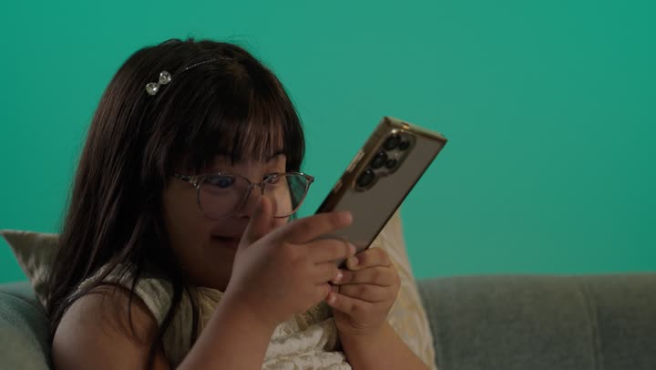 تنمية قدرات اصحاب الهمم، بورتريه لطفلة سعودية مصابة بمتلازمه داون ترتدى النظارة تجلس على الاريكة تمسك الهاتف، التعلم عن طريق الانترنت، استخدام التكنولوجيا والأجهزة التقنية الحديثة، خلفية حضراء