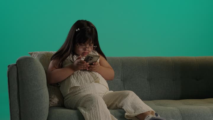 بورتريه لطفلة سعودية مصابة بمتلازمه داون ترتدى النظارة تجلس على الاريكة تمسك الهاتف، التعلم عن طريق الانترنت، استخدام التكنولوجيا والأجهزة التقنية الحديثة المتطورة، خلفية حضراء