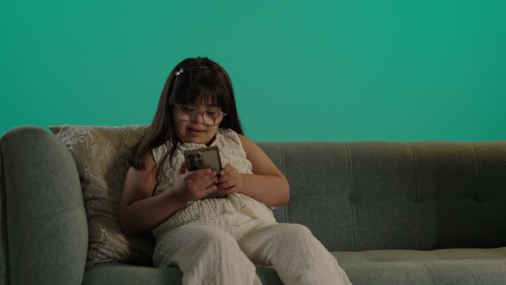 استخدام التكنولوجيا والأجهزة التقنية الحديثة المتطورة، بورتريه لطفلة سعودية مصابة بمتلازمه داون ترتدى النظارة تجلس على الاريكة تمسك الهاتف، التعلم عن طريق الانترنت، خلفية حضراء