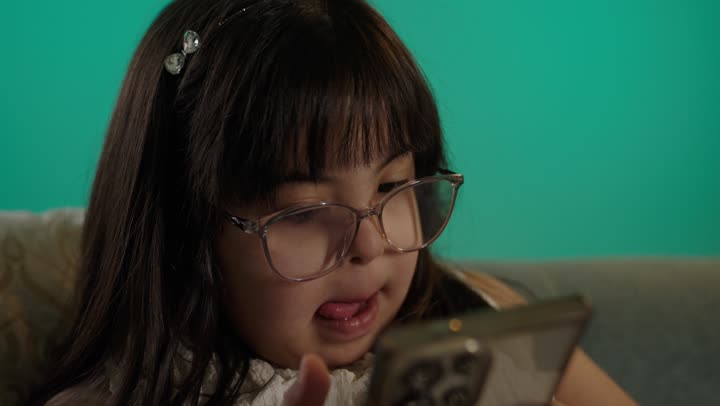 التعلم عن طريق الانترنت، بورتريه لطفلة سعودية مصابة بمتلازمه داون ترتدى النظارة تمسك الهاتف، خلفية حضراء