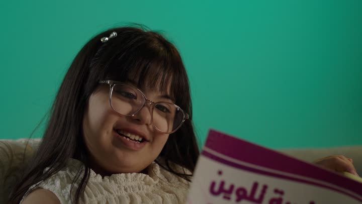 Enhancing mental activity, illustrative means for education, spending enjoyable times, a portrait of a Gulf Arab Saudi girl with Down syndrome wearing glasses sitting on the sofa holding a coloring book, green background.