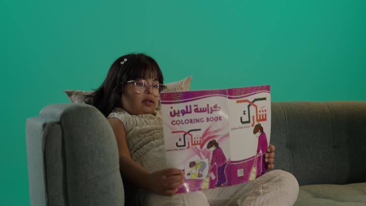 وسائل ايضاح ترفيفهيه للتعليم، تعزيز النشاط الذهني، قضاء اوقات ممتعه، بورتريه لطفلة عربية خليجية سعودية مصابة بمتلازمه داون ترتدى نظارة تجلس على الاريكة تمسك كراسة التلوين، خلفية خضراء