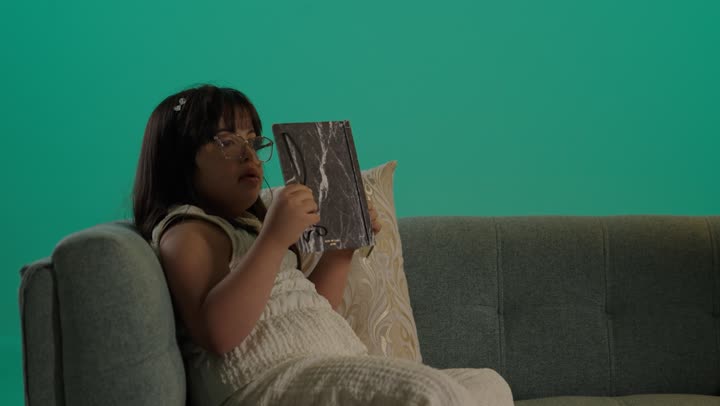 مفهوم هوايه المطالعه، قضاء اوقات ممتعه، بورتريه لطفلة عربية خليجية سعودية مصابة بمتلازمه داون ترتدى نظارة تجلس على الاريكة تمسك كتاب، خلفية خضراء