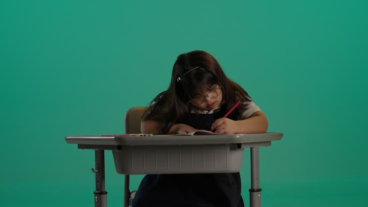 مفهوم الاهتمام بالهوايات، بورتريه لطفلة سعودية مصابة بمتلازمه داون ترتدى النظارة تجلس على   الطاولة تقوم بالتلوين، خلفية حضراء