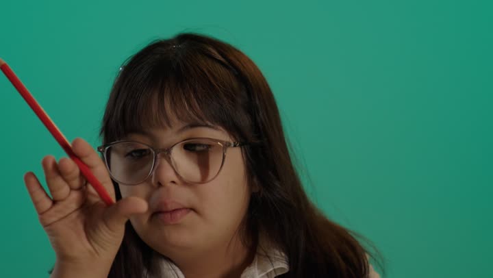 Holding a coloring pencil, the concept of caring for hobbies, a portrait of a Saudi girl with Down syndrome wearing glasses sitting at a table coloring, green background.