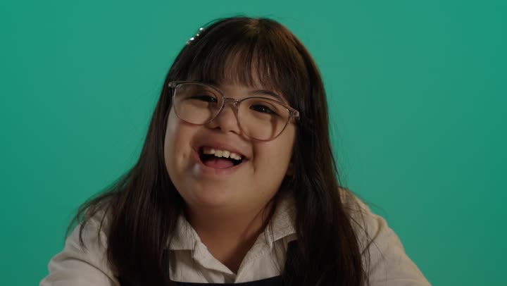 A portrait of a Saudi girl with Down syndrome smiling and looking at the camera while coloring, caring for and developing children's skills, having a good time, with a green background.