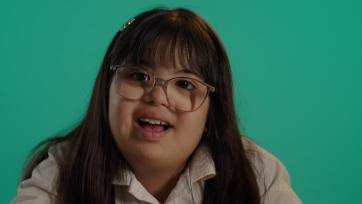 ايماءات السرور و السعاده، بورتريه لطفلة سعودية مصابة بمتلازمه داون مبتسمة  ترتدى نظارة تنظر الى الكاميرا، رعاية و تطوير مهارات الأطفال، قضاء وقت ممتع، خلفية خضراء