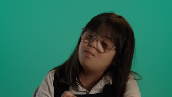 Inspiration for drawing from surrounding objects, a close-up portrait of the hand of a Saudi girl with Down syndrome wearing glasses, holding colors, sitting at a table looking at the camera, nurturing talent and interest in Saudi Arabia, green background.