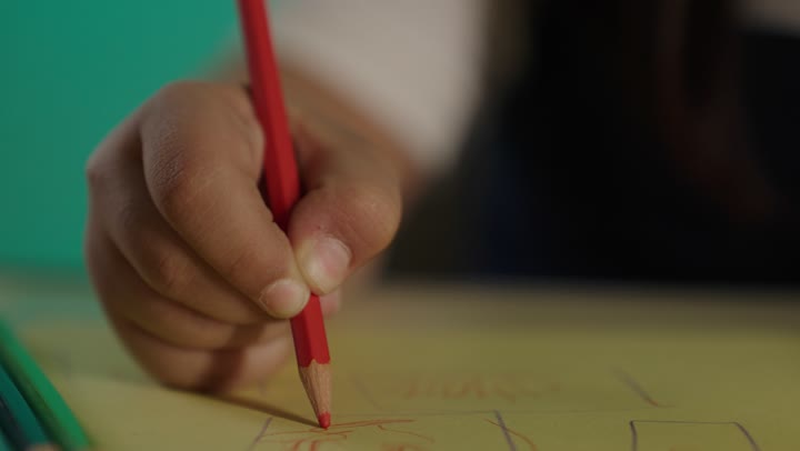 A close-up portrait of the hand of a Saudi girl with Down syndrome wearing glasses, holding colors, sitting at a table. Nurturing talent and caring for it in Saudi Arabia, developing artistic skills for people with determination, with a green background.