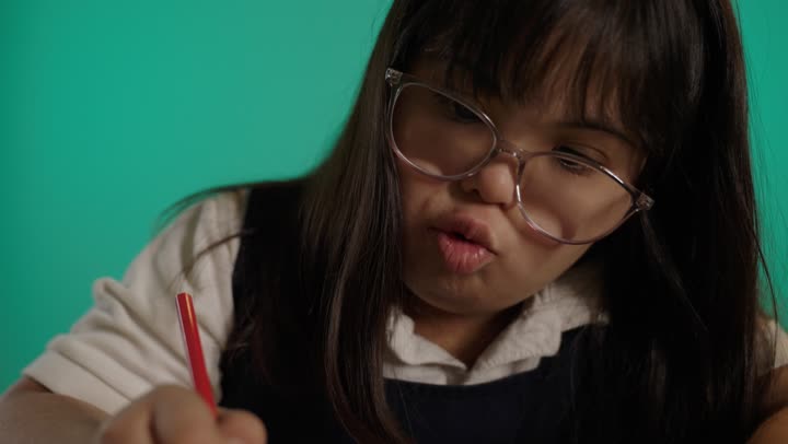 رعاية الموهبة والاهتمام بها فى السعودية, تنمية المهارة الفنية لذوي الهمم، بورتريه لطفلة سعودية مصابة بمتلازمه داون ترتدى النظارة تمسك الالوان تجلس على الطاولة، خلفية حضراء