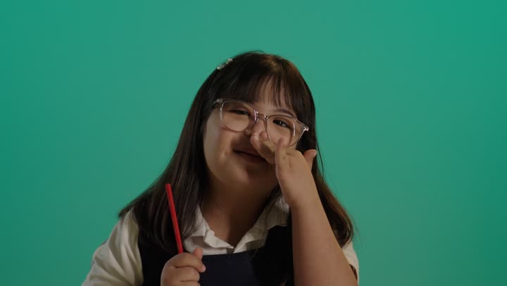 استخدام أقلام التلوين الخشبية, تنمية المهارة الفنية لذوي الهمم، بورتريه لطفلة سعودية مصابة بمتلازمه داون ترتدى النظارة تمسك الالوان تنظر الى الكاميرا، خلفية حضراء