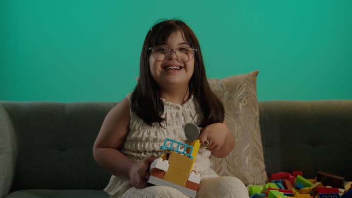 مفهوم الاهتمام بالهوايات، قضاء اوقات ممتعه فى المنزل، تنمية المهارات الذهنيه والفكريه، تعابير السعاده والسرور، بورتريه لطفلة سعودية مصابة بمتلازمه داون تجلس على الاريكة تقوم باللعب بالمكعبات, خلفية خضراء