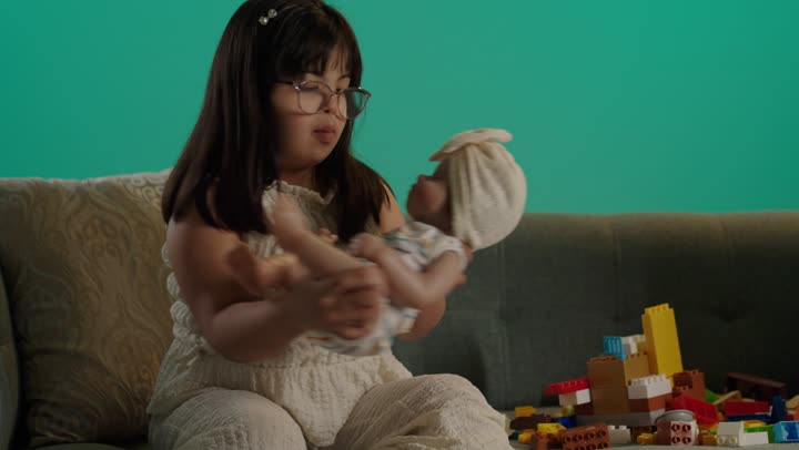 اللعب واللهو والمرح بحماس ونشاط، بورتريه لطفلة سعودية مصابة بمتلازمه داون ترتدى النظارة تلعب بالدمية، قضاء وقت ممتع بالالعاب، خلفية حضراء