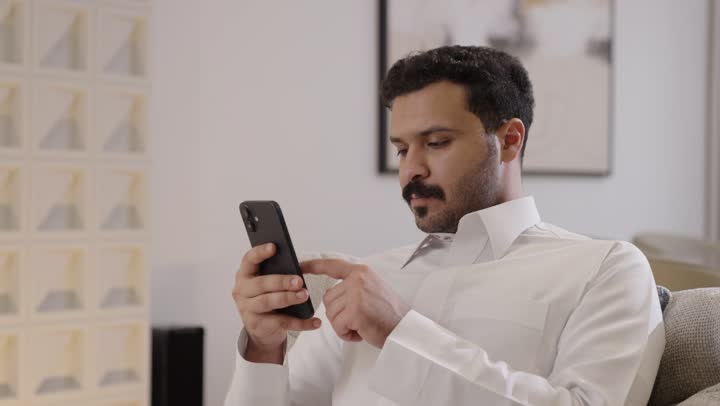 الدردشة عبر الانترنت، رجل عربي خليجي سعودي مبتسم يرتدي الثوب التقليدي يمسك الجوال بيده يجلس على الاريكة، تصفح وسائل التواصل الاجتماعي