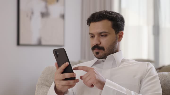 Using a mobile phone to accomplish tasks, a smiling Arab Gulf Saudi man wearing traditional attire holds a mobile phone in his hand while sitting on the sofa, browsing social media.