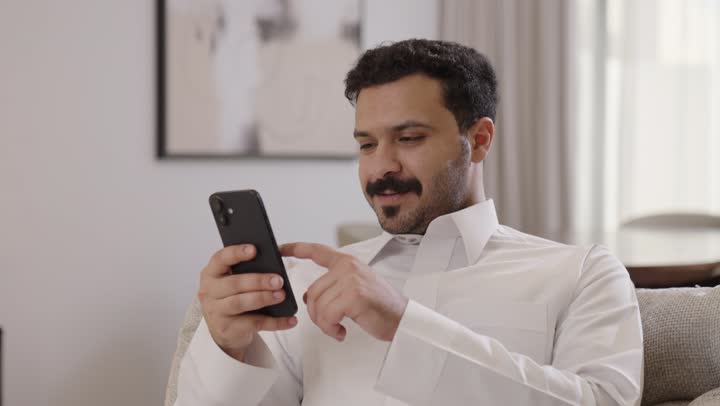 مفهوم الجلوس والراحة، رجل عربي خليجي سعودي مبتسم يرتدي الثوب التقليدي يمسك الهاتف المحمول بيده ينظر الى الكاميرا، تصفح وسائل التواصل الاجتماعي عبر الهاتف